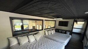 Una cama doble con cubrecama acolchado, un espejo grande encima y un televisor montado en la pared.