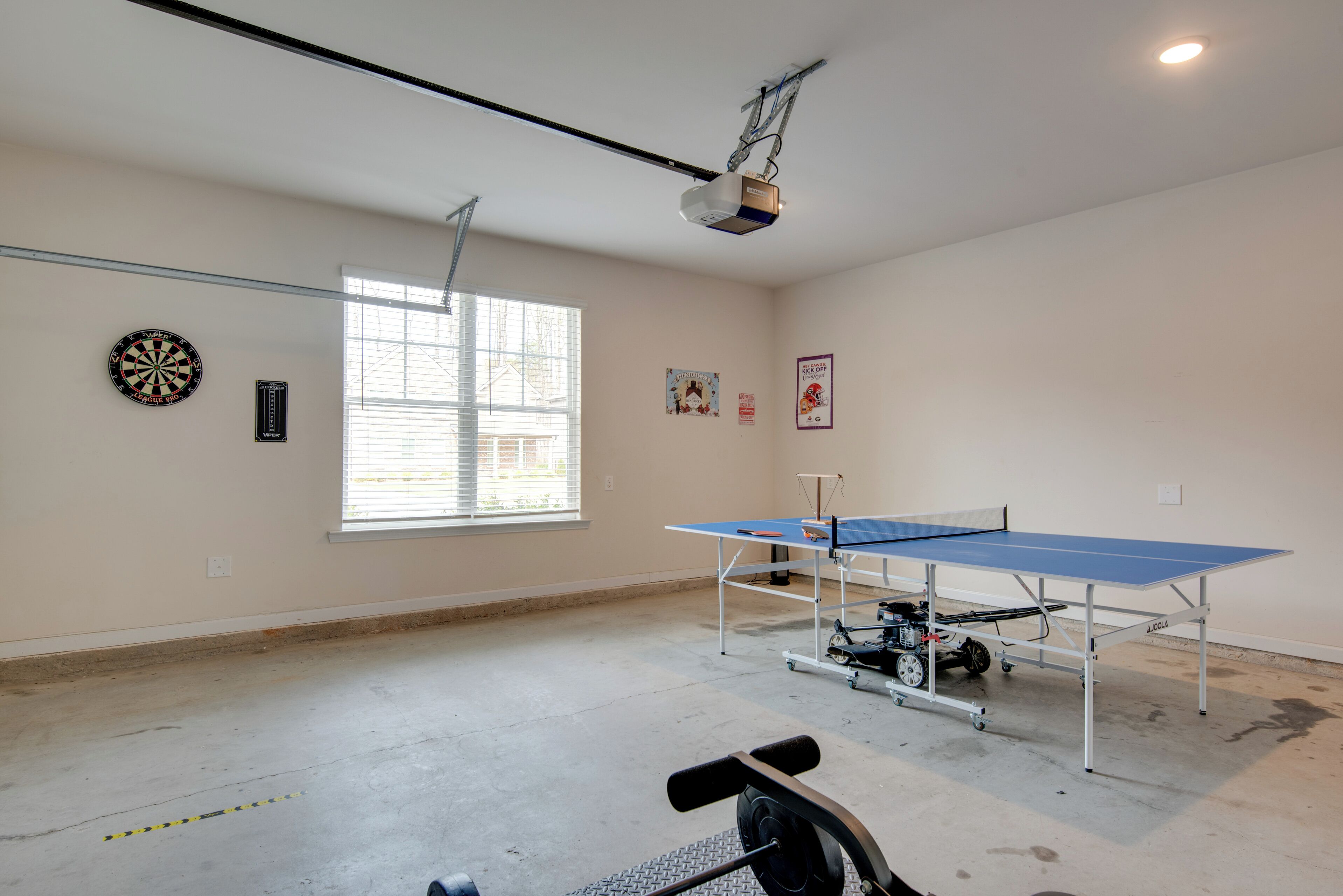 Una habitación con mesa de ping pong, proyector en el techo, dardo y ventana con persianas.