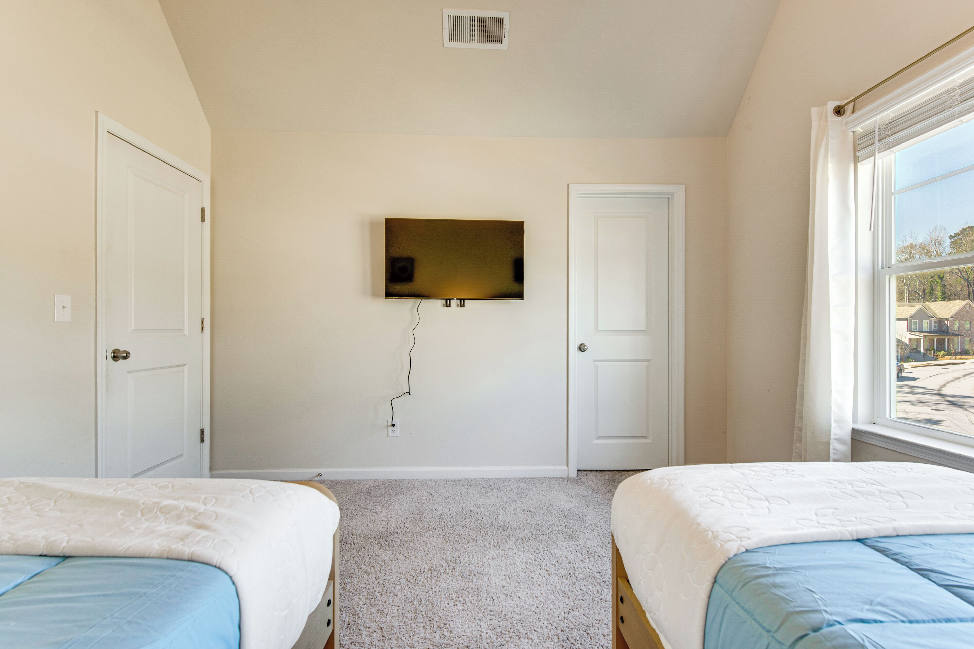 A bedroom with two beds, a television mounted on the wall, and a window with a view of the outside.