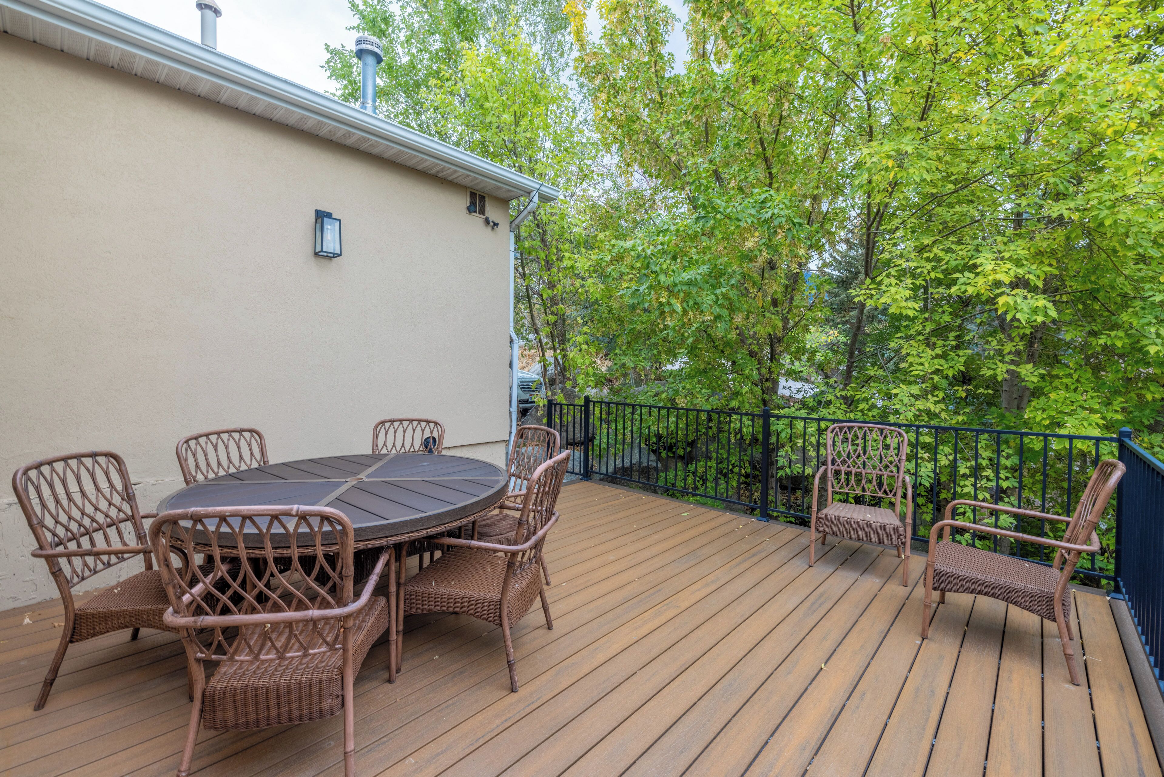 A wooden deck with a round table and wicker chairs, surrounded by a metal railing and trees.
