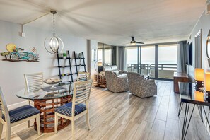 Ein modernes Wohnzimmer mit einem Glastisch, gemütlicher Sitzgelegenheit, einem Deckenventilator und einem großen Fenster mit Aussicht.