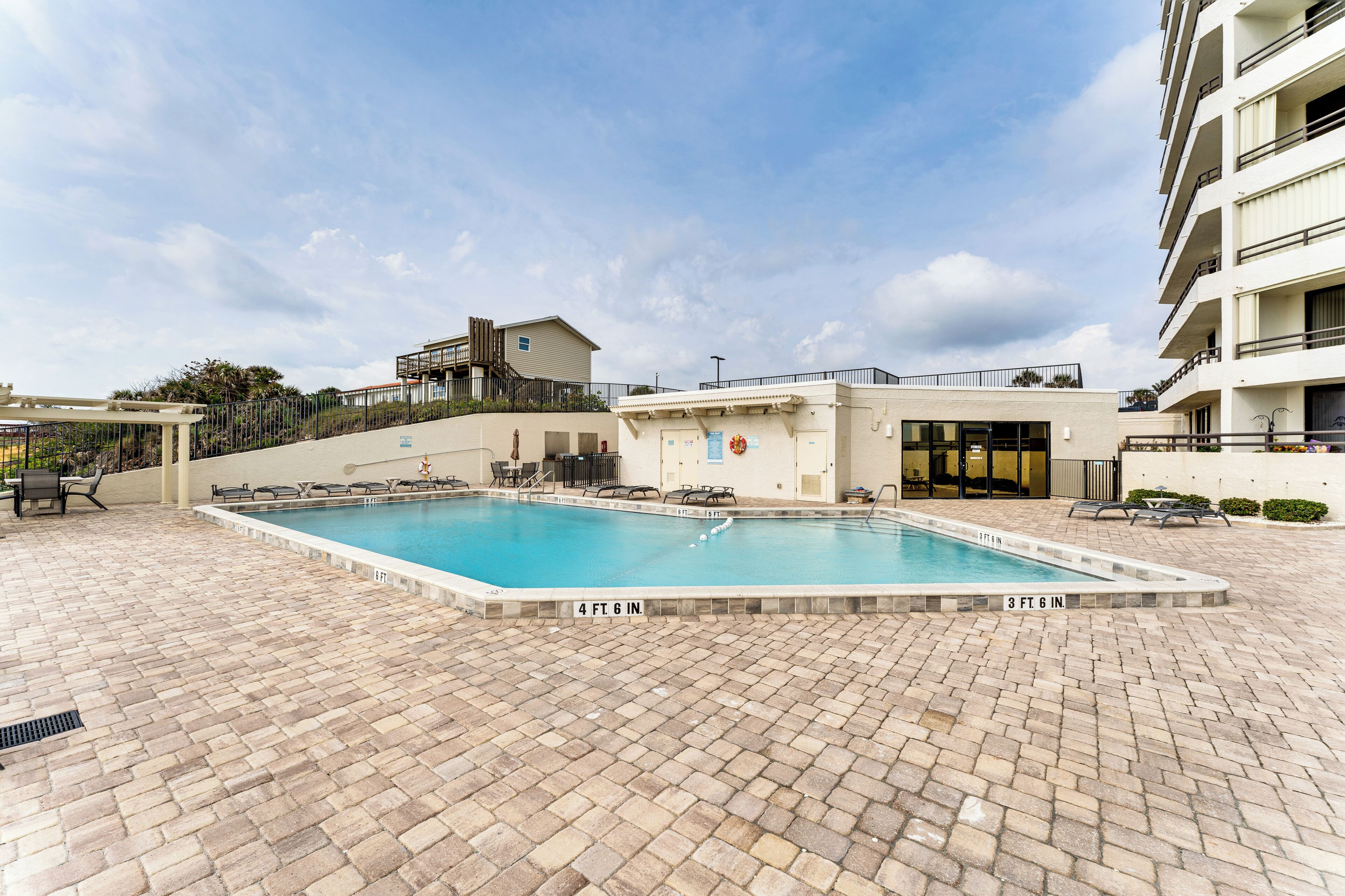 A swimming pool with a brick patio, a building with a balcony, and a staircase leading to a higher level.