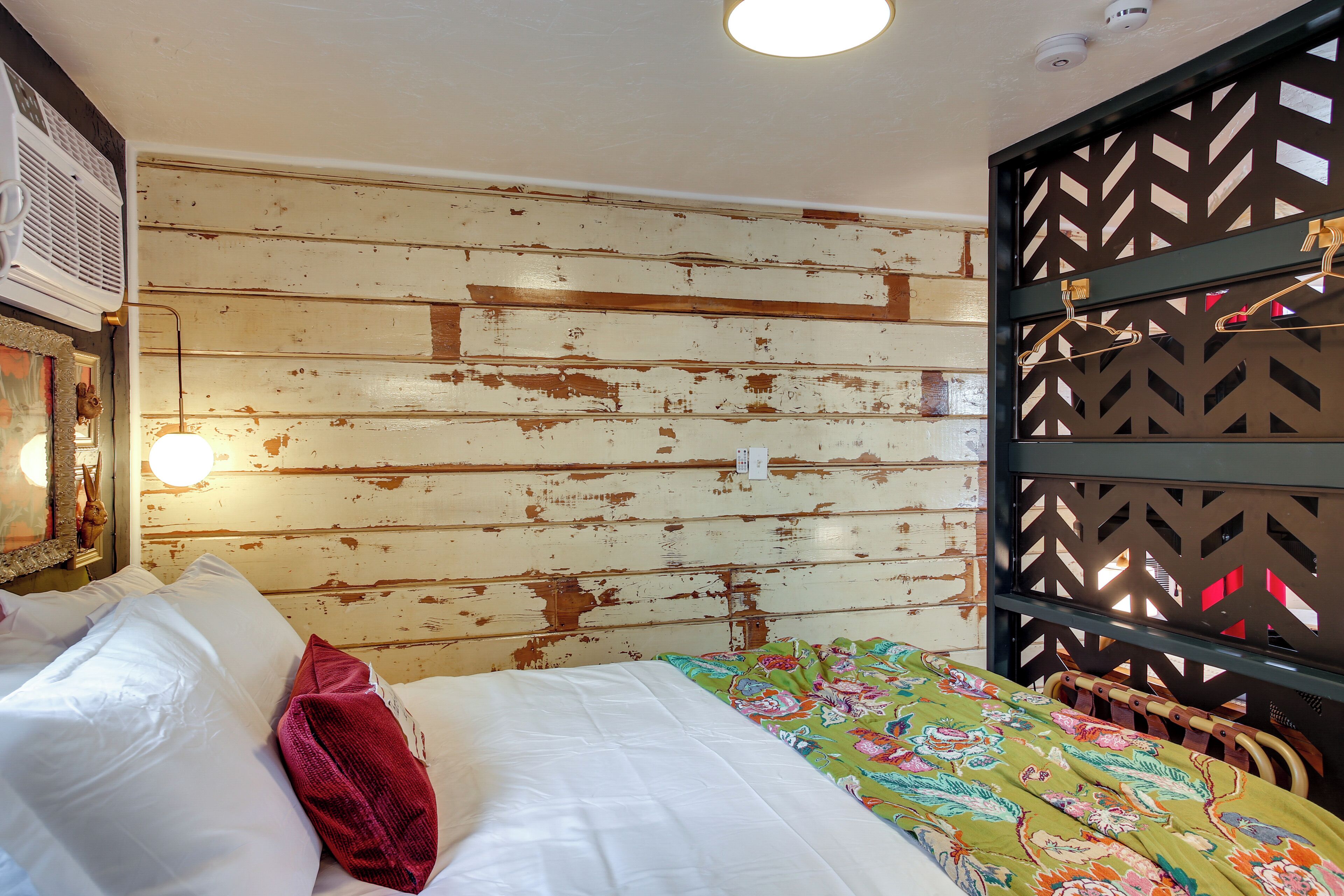 A room with a bed, a wooden wall with peeling paint, a hanging light fixture, and a decorative panel with a geometric pattern.