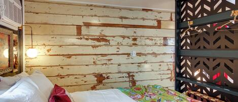 A room with a bed, a wooden wall with peeling paint, a hanging light fixture, and a decorative panel with a geometric pattern.