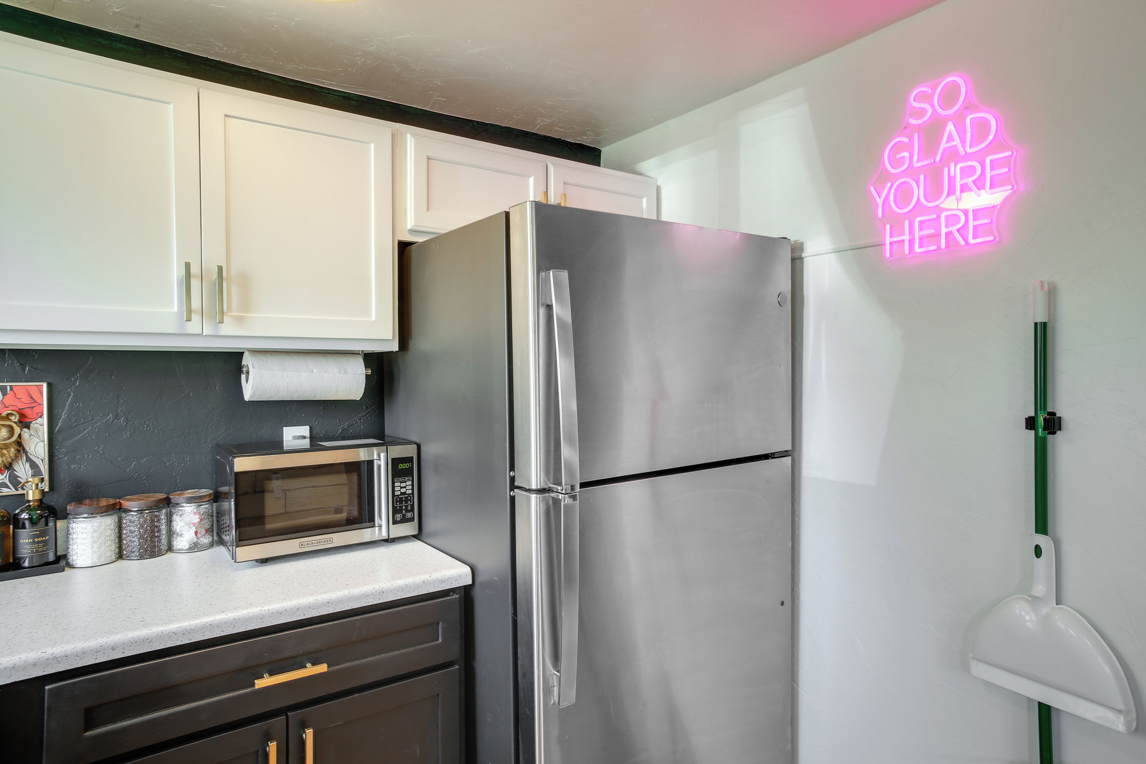 A kitchen with white cabinets, a stainless steel refrigerator, a microwave, and a neon sign on the wall.