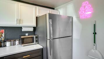 A kitchen with white cabinets, a stainless steel refrigerator, a microwave, and a neon sign on the wall.
