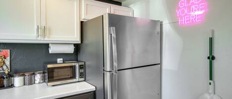 A kitchen with white cabinets, a stainless steel refrigerator, a microwave, and a neon sign on the wall.
