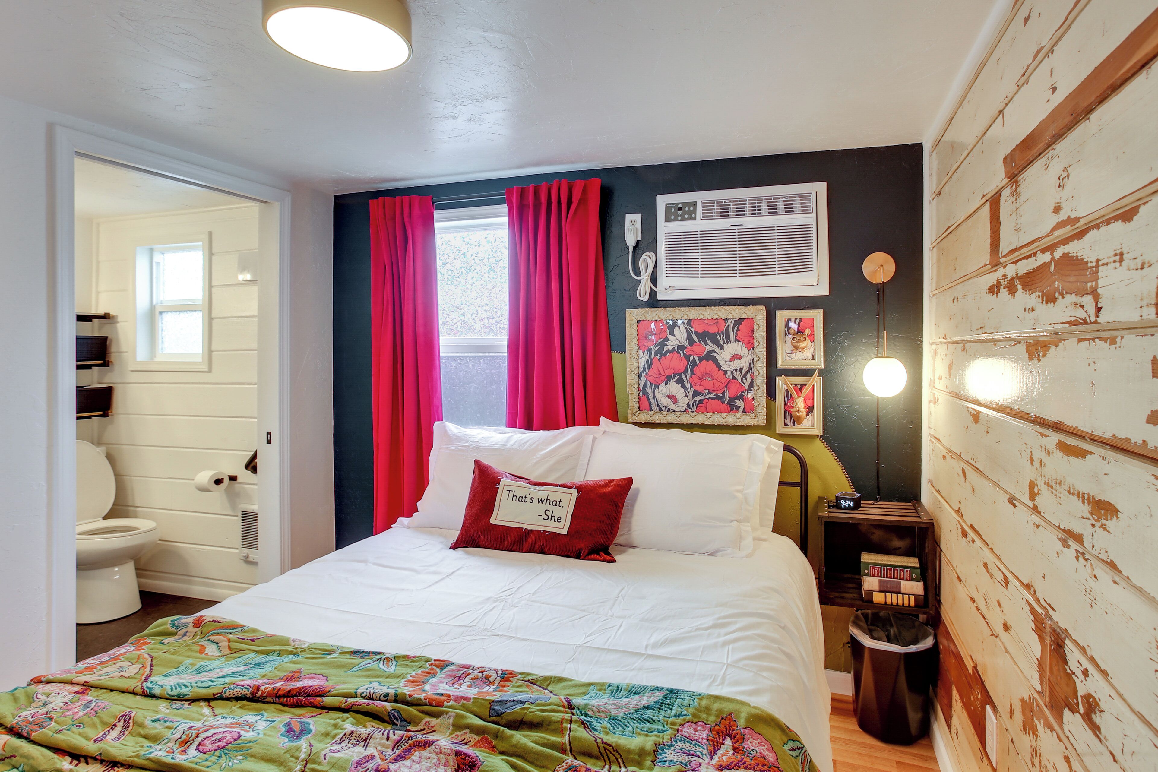 Een slaapkamer met een bed, een raam met gordijnen, een wandairconditioner, een nachtkastje met lamp en een prullenbak.