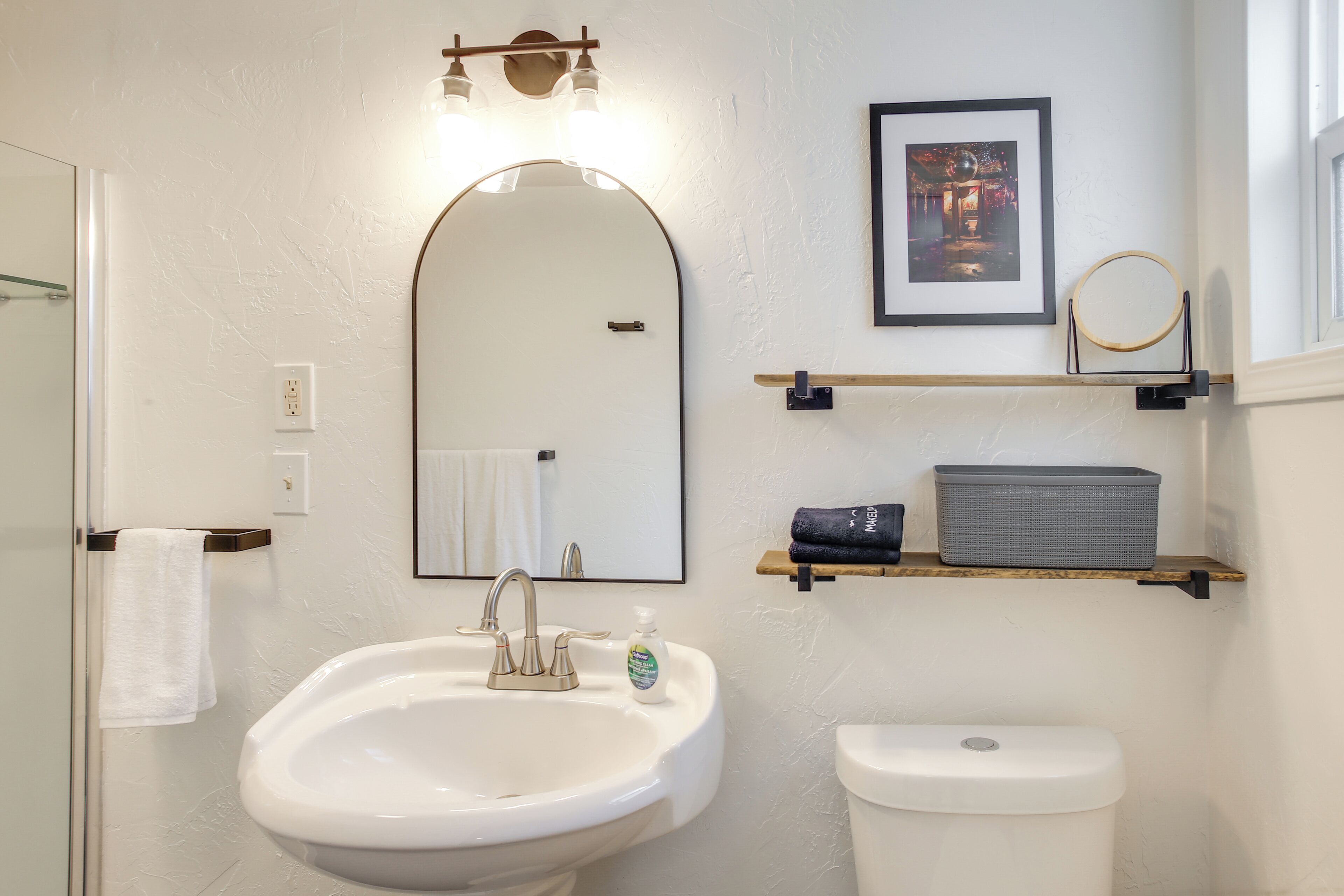 A bathroom with a white sink, a mirror, a toilet, and a shelf with towels and toiletries.