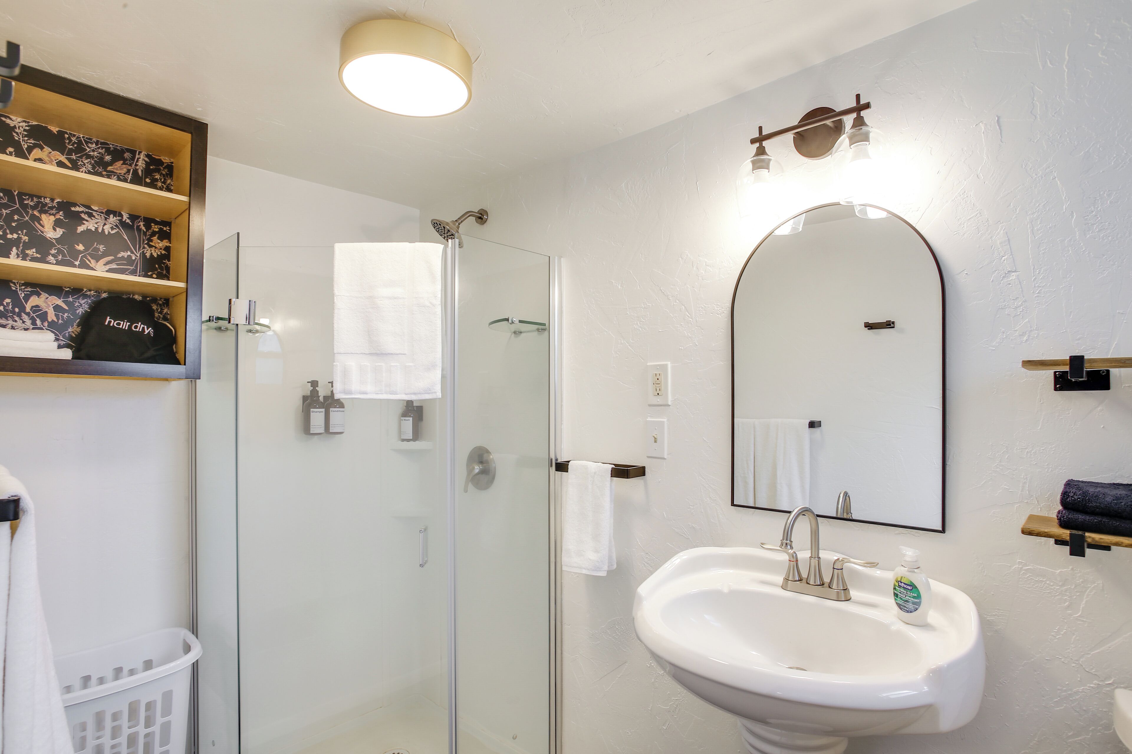Un baño con cabina de ducha de vidrio, un lavamanos blanco, un espejo y un estante con artículos de aseo.