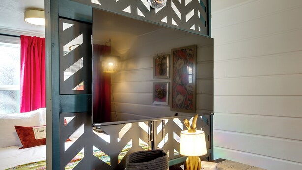 Una habitación con cama, escritorio con lámpara, espejo y un panel decorativo en la pared.