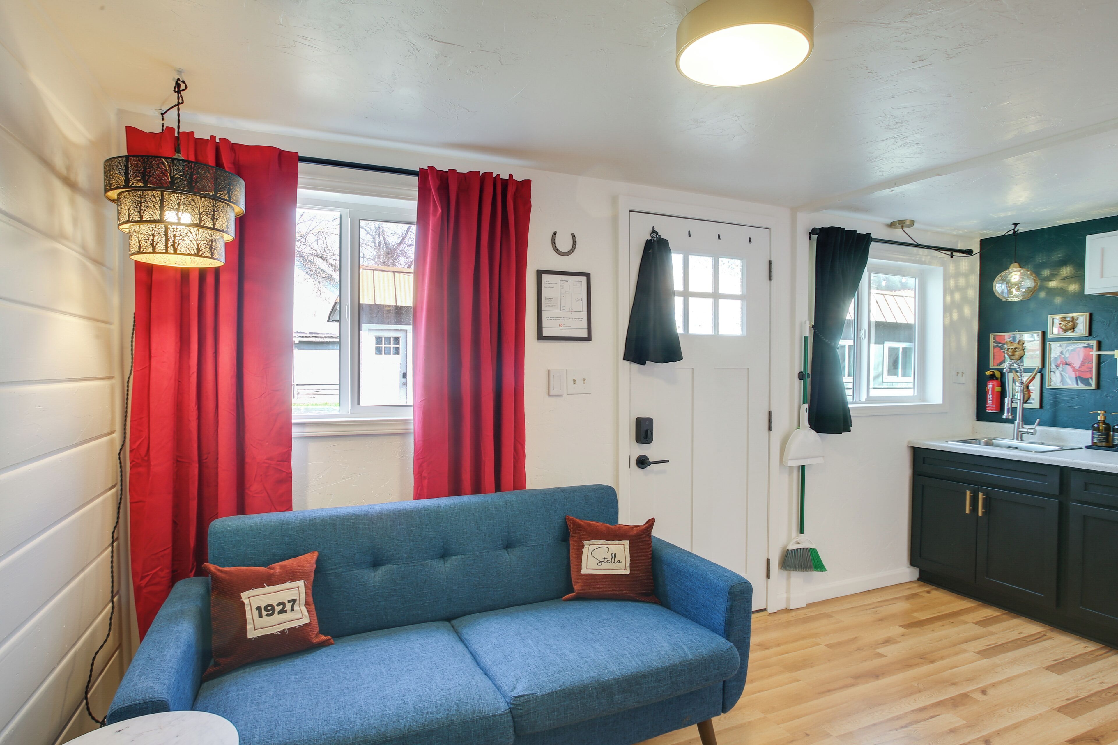 A room with a blue sofa, red curtains, a white door, a kitchen area, and a hanging light fixture.