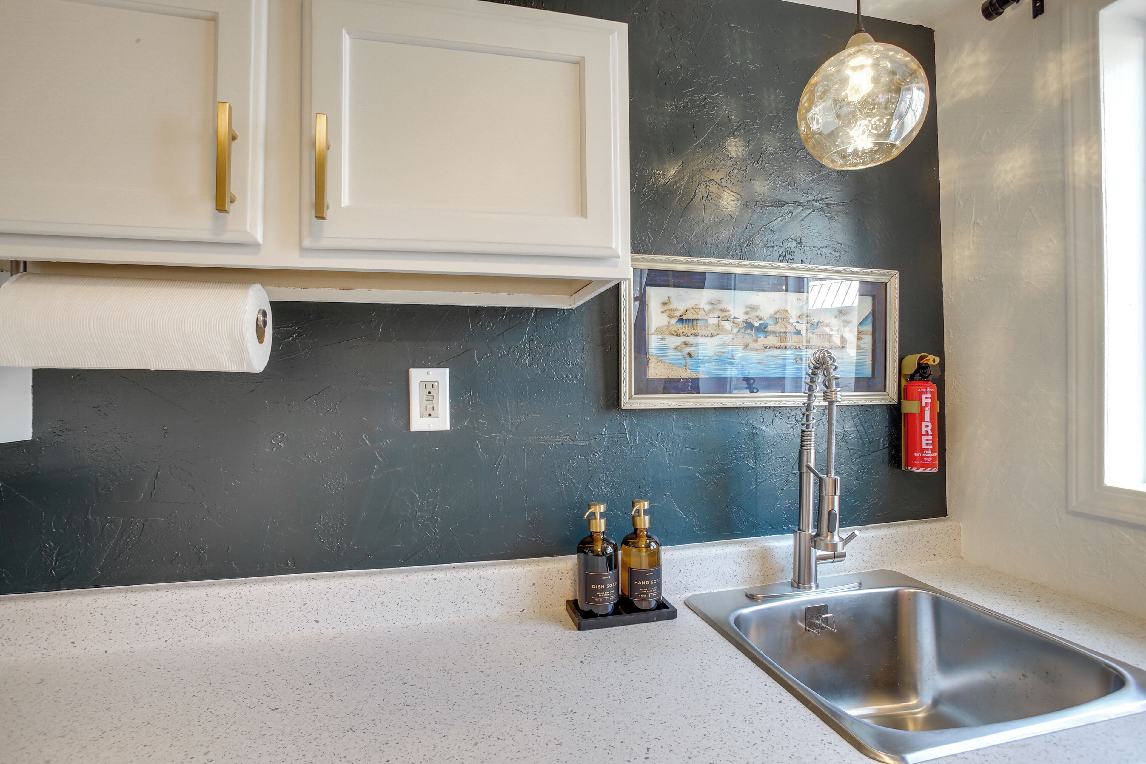 A kitchen with white cabinets, a black countertop, a stainless steel sink, a hanging light fixture, and two hand sanitizer bottles.