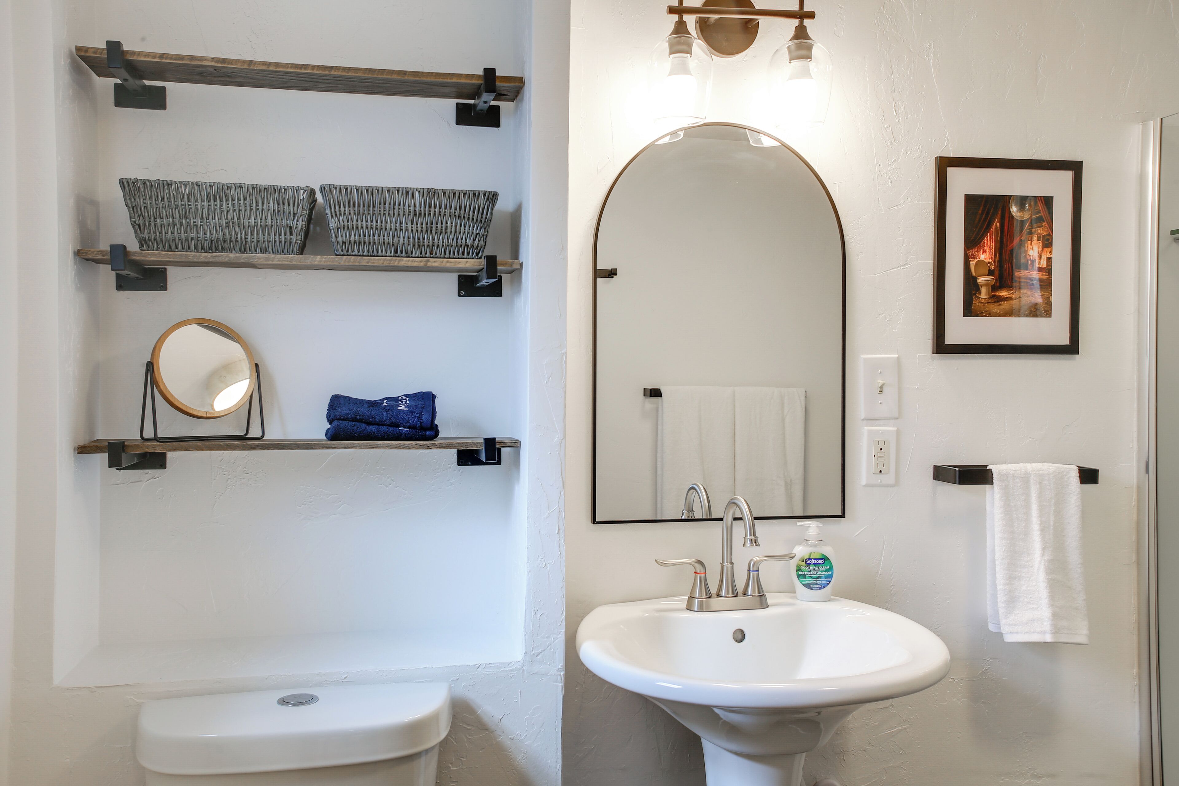 A bathroom with a toilet, a sink, a mirror, shelves with baskets, and a framed picture on the wall.