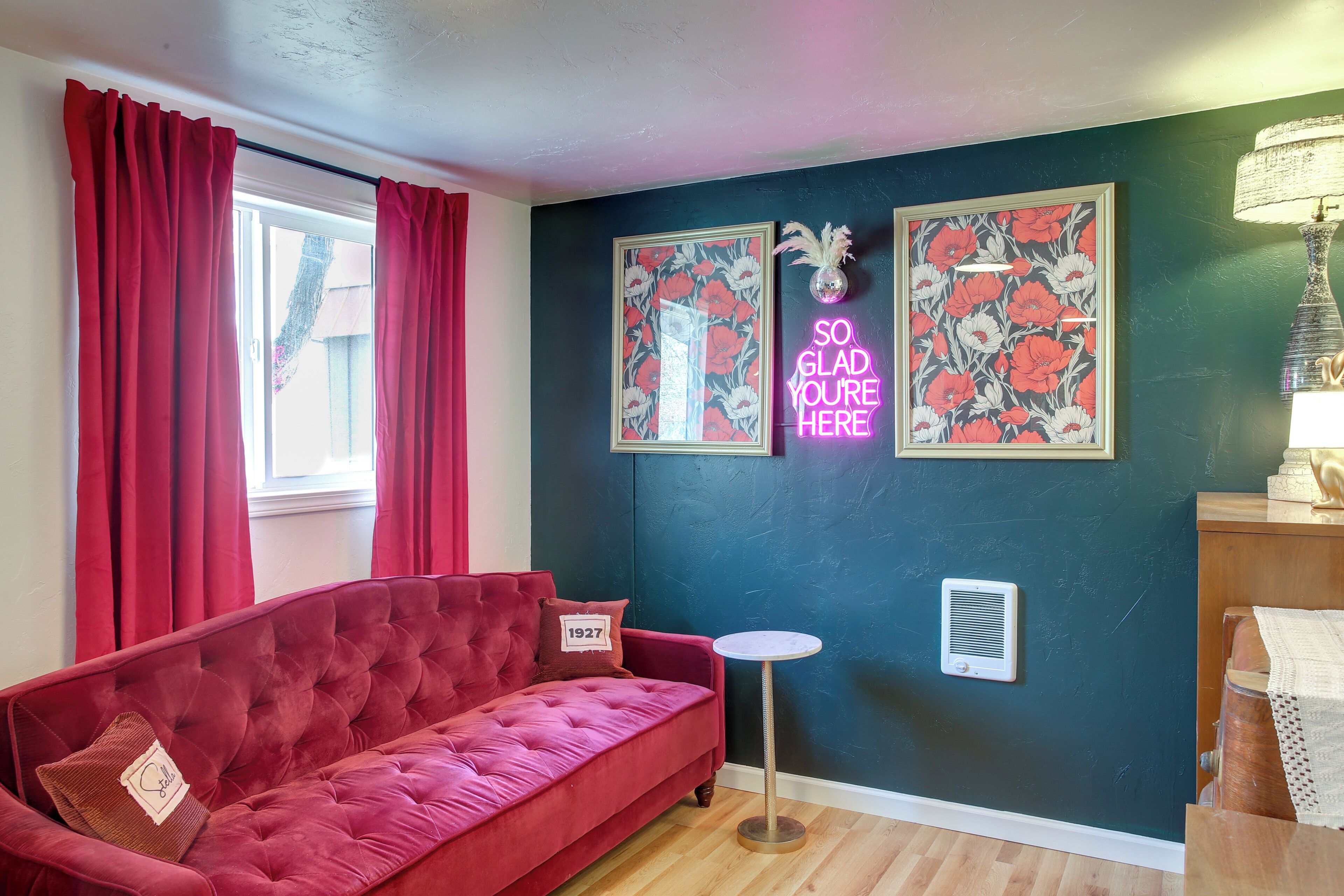 A room with a red velvet sofa, a small round table, a wall-mounted air vent, and two framed floral pictures.