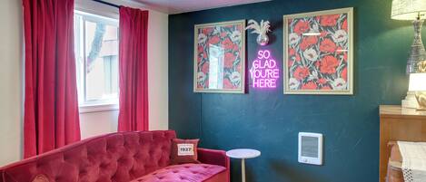 A room with a red velvet sofa, a small round table, a wall-mounted air vent, and two framed floral pictures.