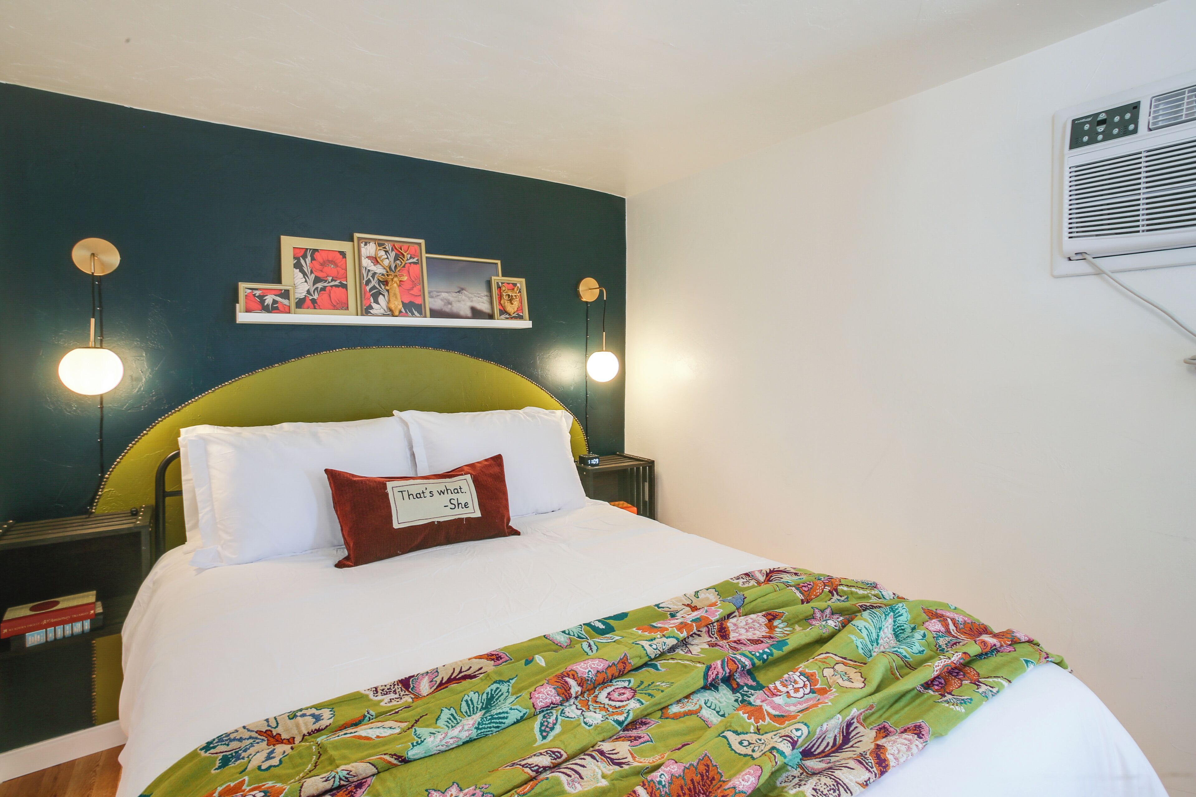 A neatly made bed with a floral bedspread, a green upholstered headboard, and a wall-mounted air conditioner.