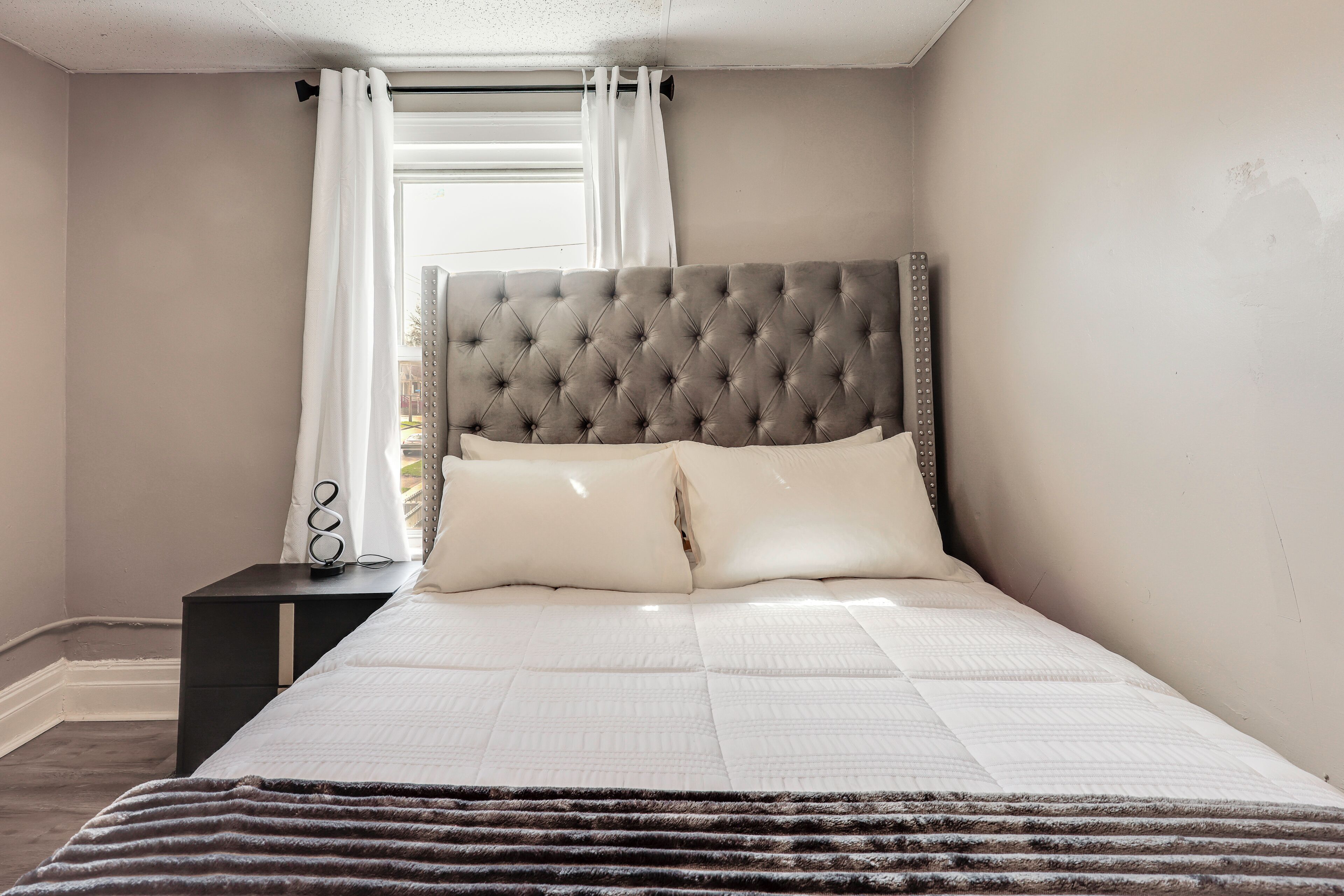 A bedroom with a tufted headboard, a bed with white bedding, a nightstand with a lamp, and a window with curtains.