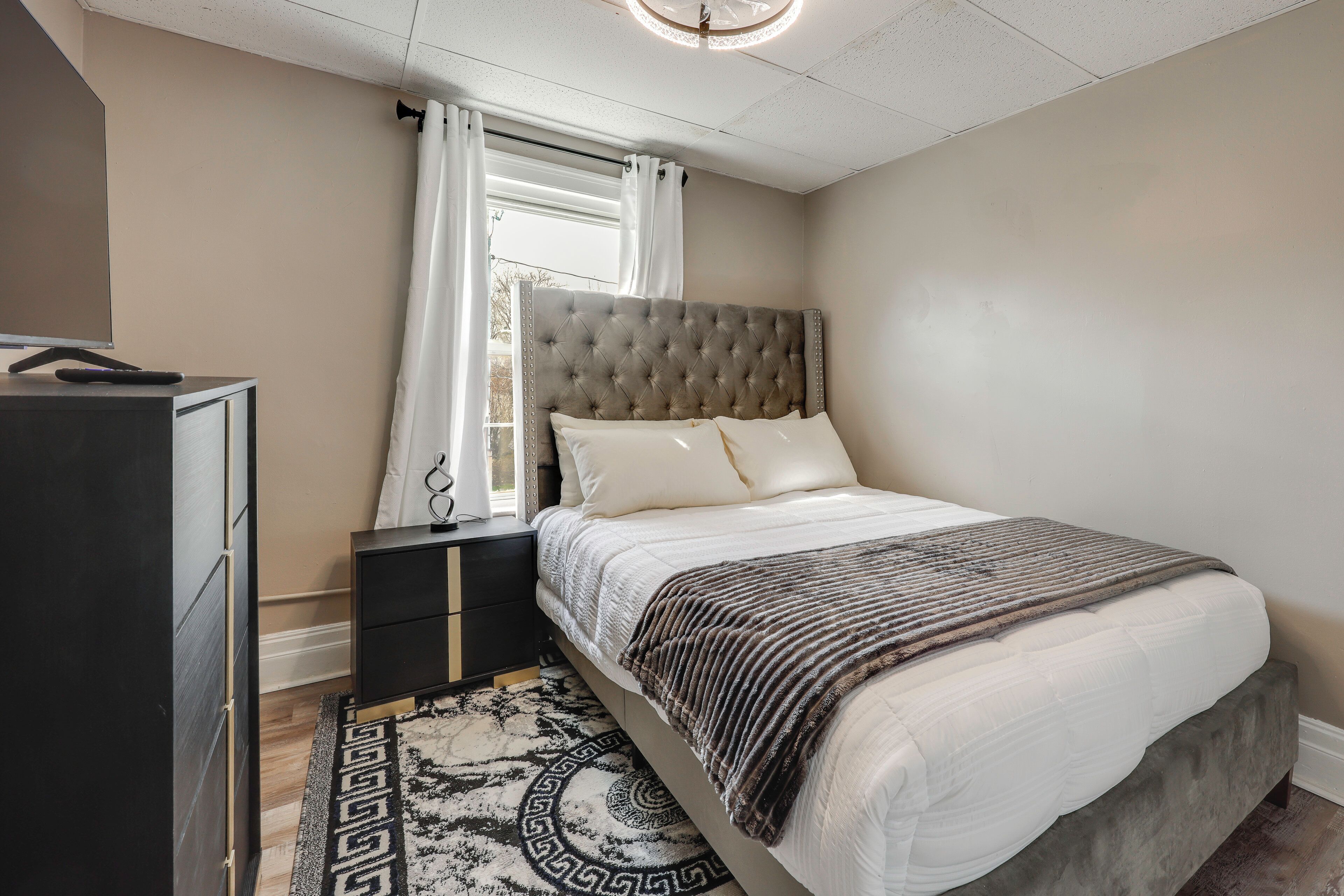 Een slaapkamer met een bed, nachtkastje, dressoir en een raam met gordijnen.