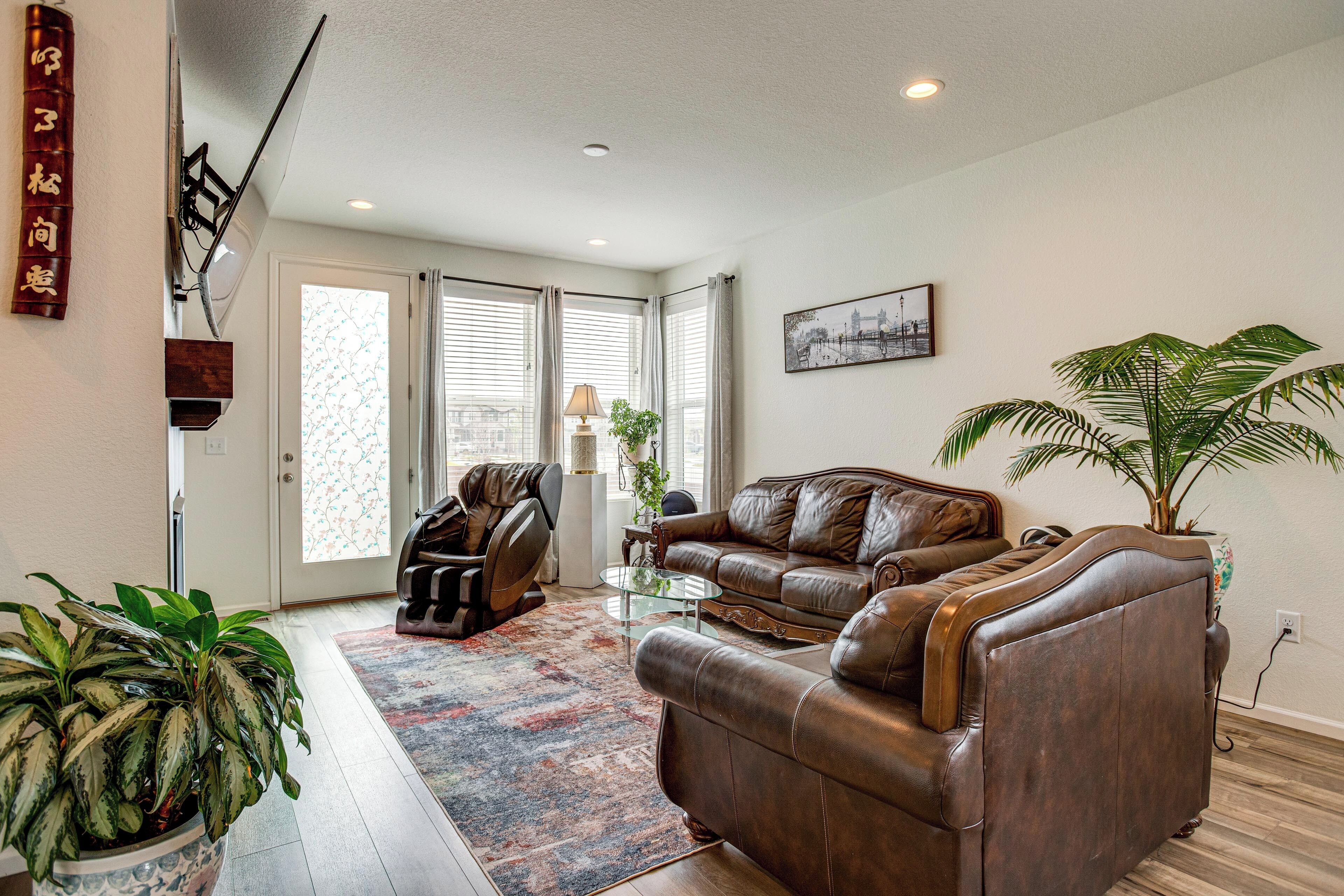 A living room with a brown leather sofa, a glass coffee table, a patterned rug, a potted plant, and a cityscape painting on the wall.