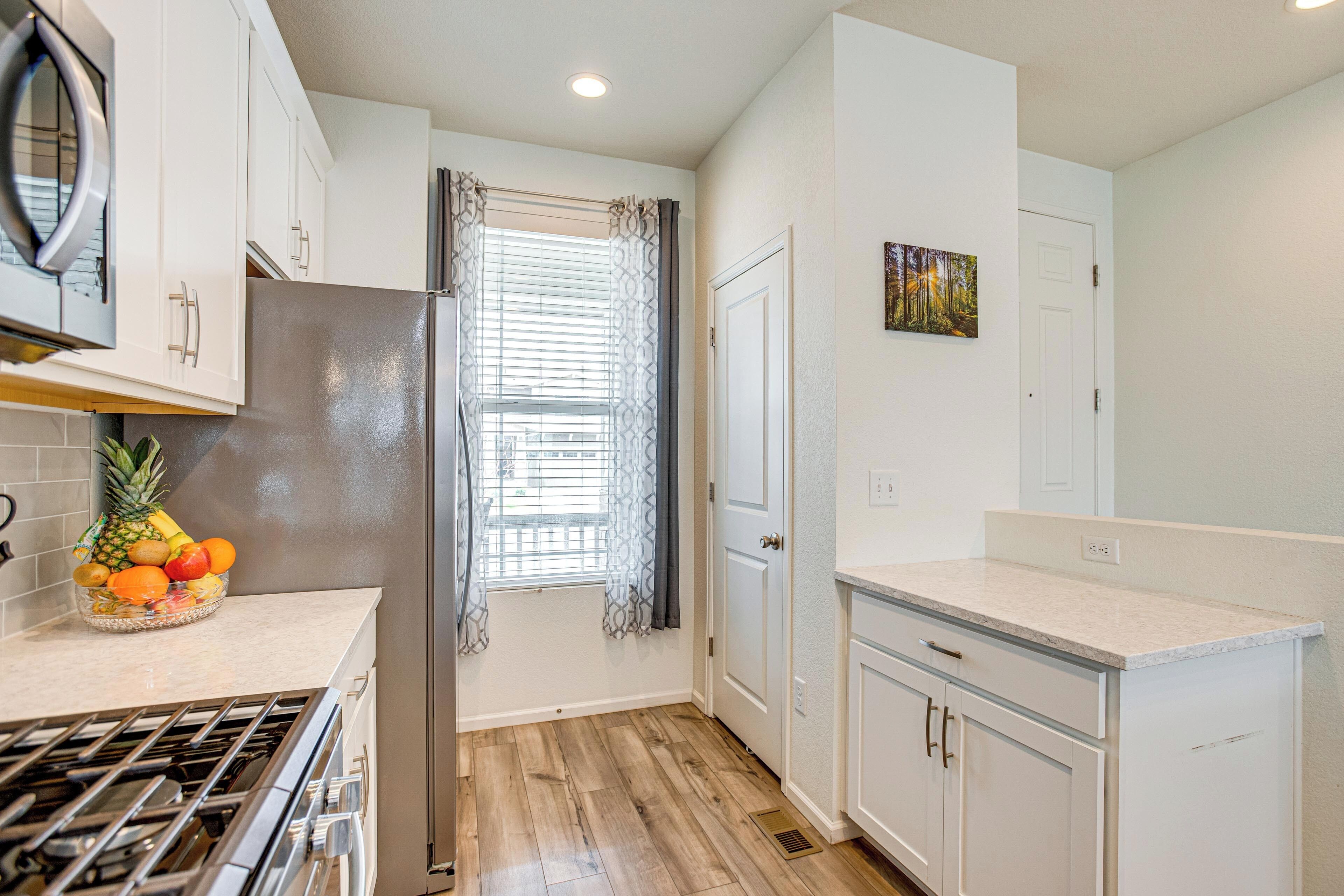 A kitchen with white cabinets, a stainless steel refrigerator, a fruit bowl, and a window with curtains.