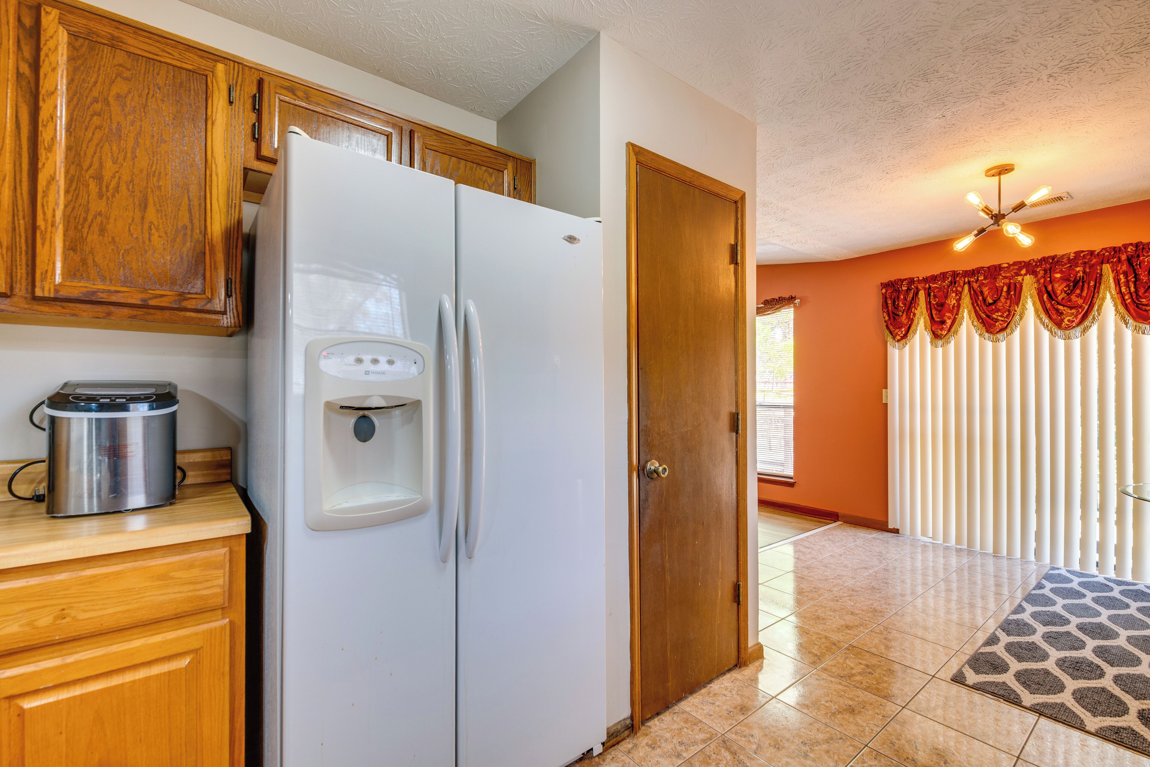 A kitchen with wooden cabinets, a white refrigerator, a coffee maker, and a door leading to a room with a window and curtains.