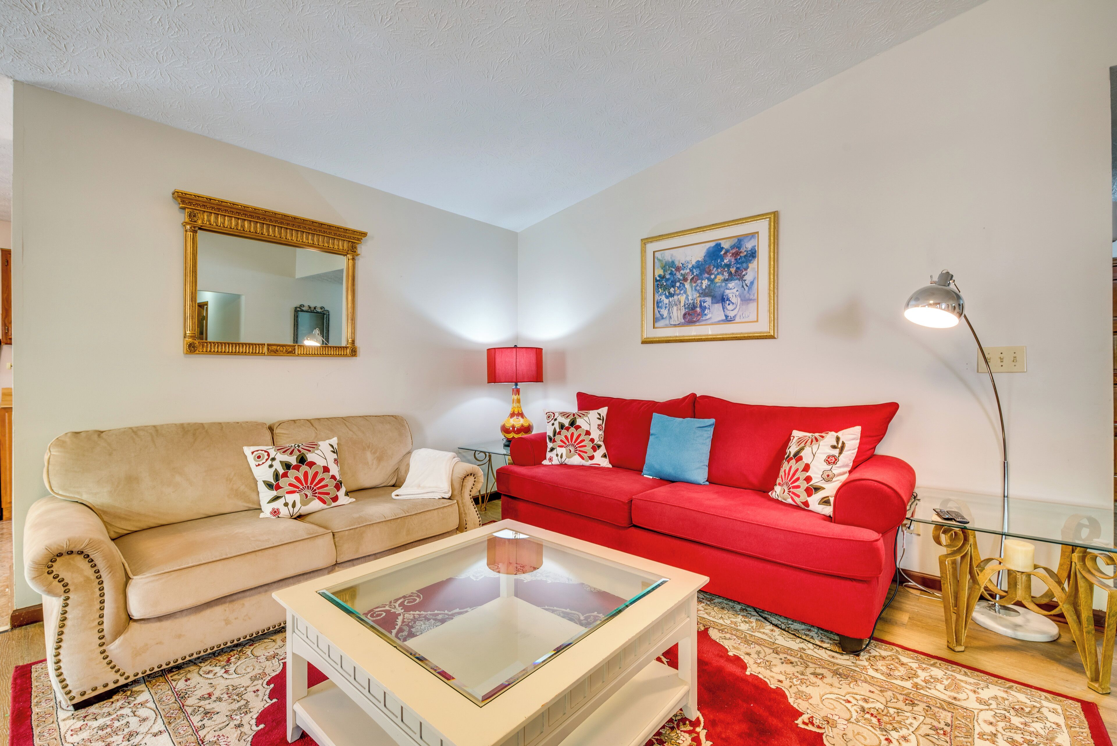 A living room with a beige sofa, a red sofa, a glass coffee table, a red lamp, a framed painting, and a mirror on the wall.