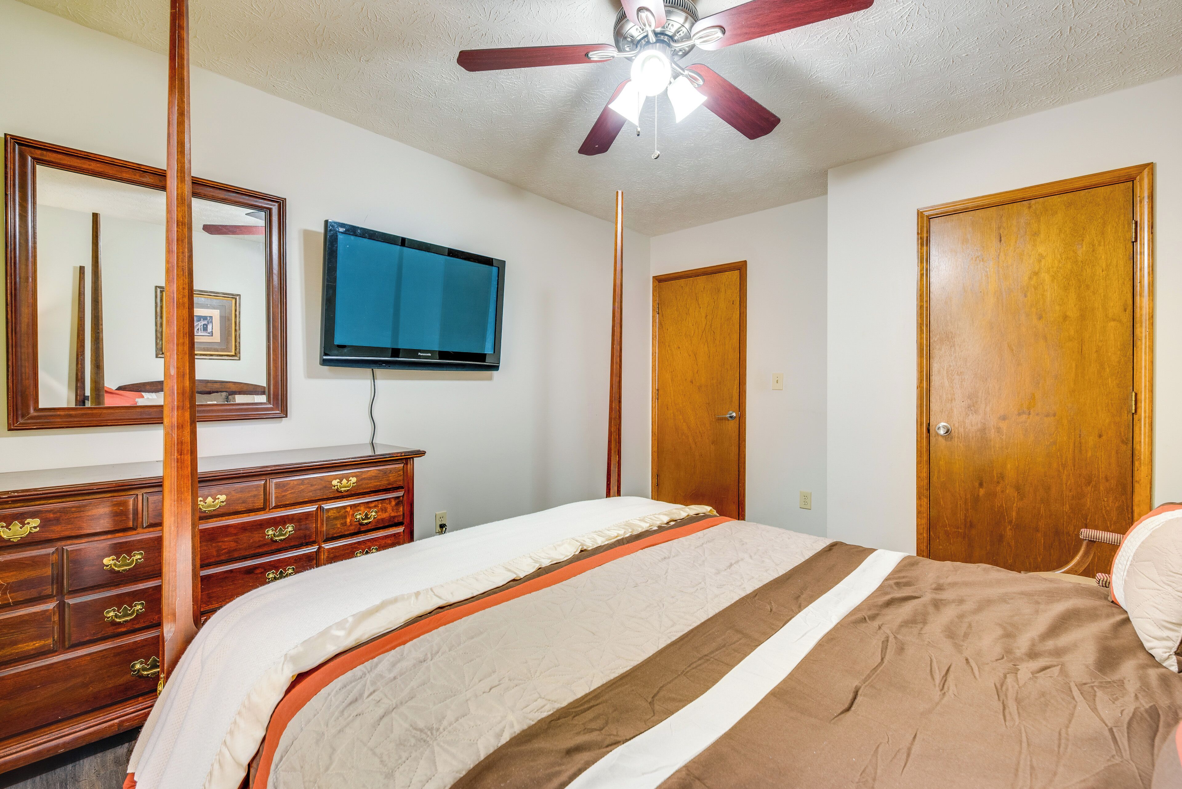 Une chambre à coucher comprenant un lit, une commode, un téléviseur, un ventilateur de plafond et des portes en bois.