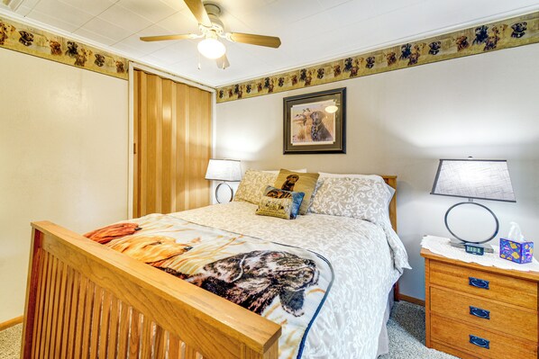 A bedroom with a wooden bed, a ceiling fan, a bedside table with a lamp, a framed picture on the wall, and a nightstand with a tissue box.