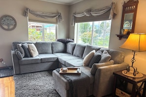 A living room with a grey sectional sofa, a small ottoman, a tray with a remote, and a lamp on a side table. - Walk to Twilight Sites! Forks Home w/ Hot Tub (Forks)