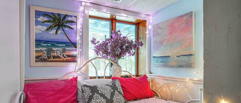 Una cama con ropa de cama estampada, un florero con flores y decoración en la pared que incluye una escena de playa y un atardecer.