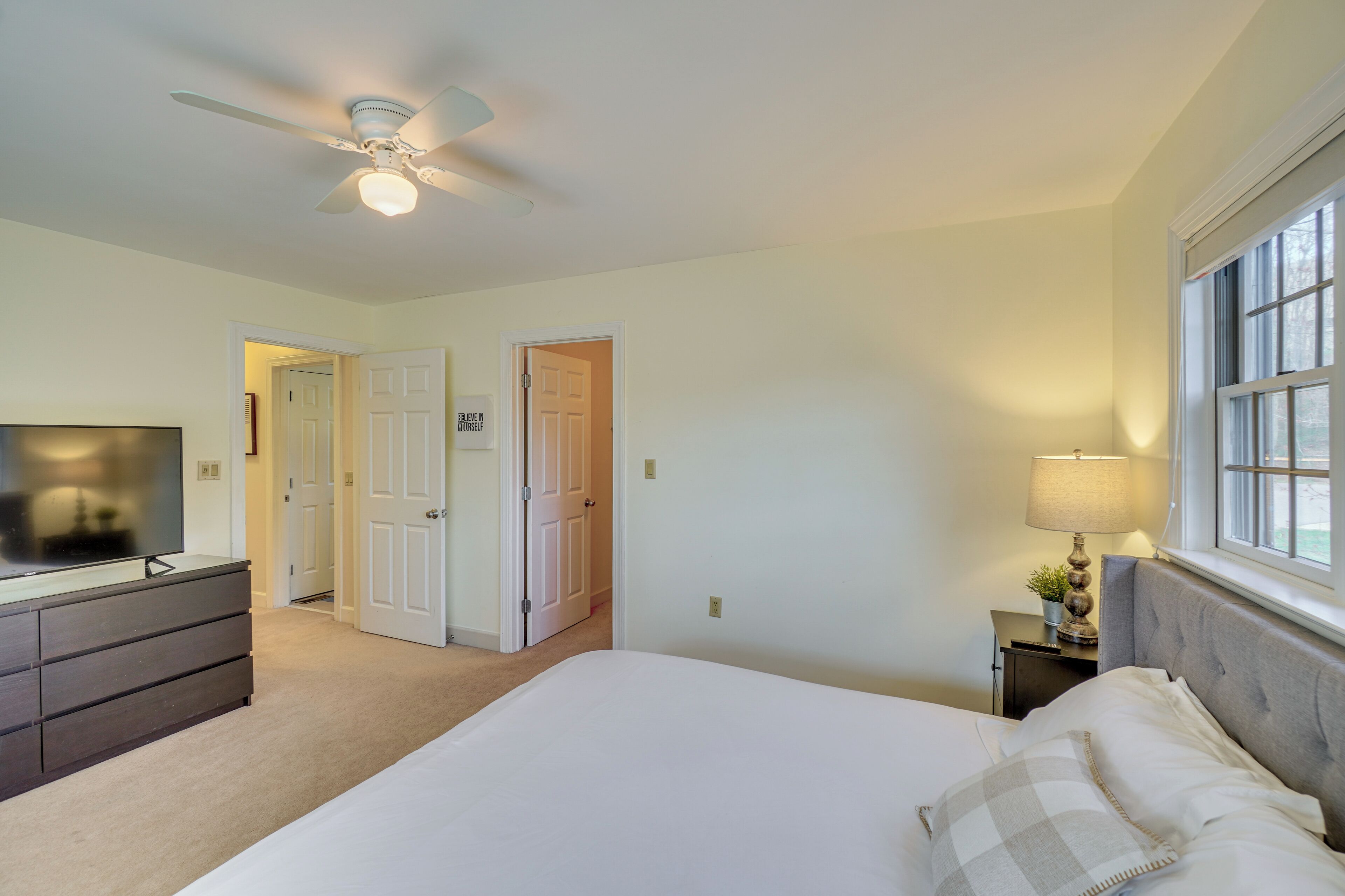 A bedroom with a bed, a dresser, a television, a ceiling fan, a nightstand with a lamp, and a window.