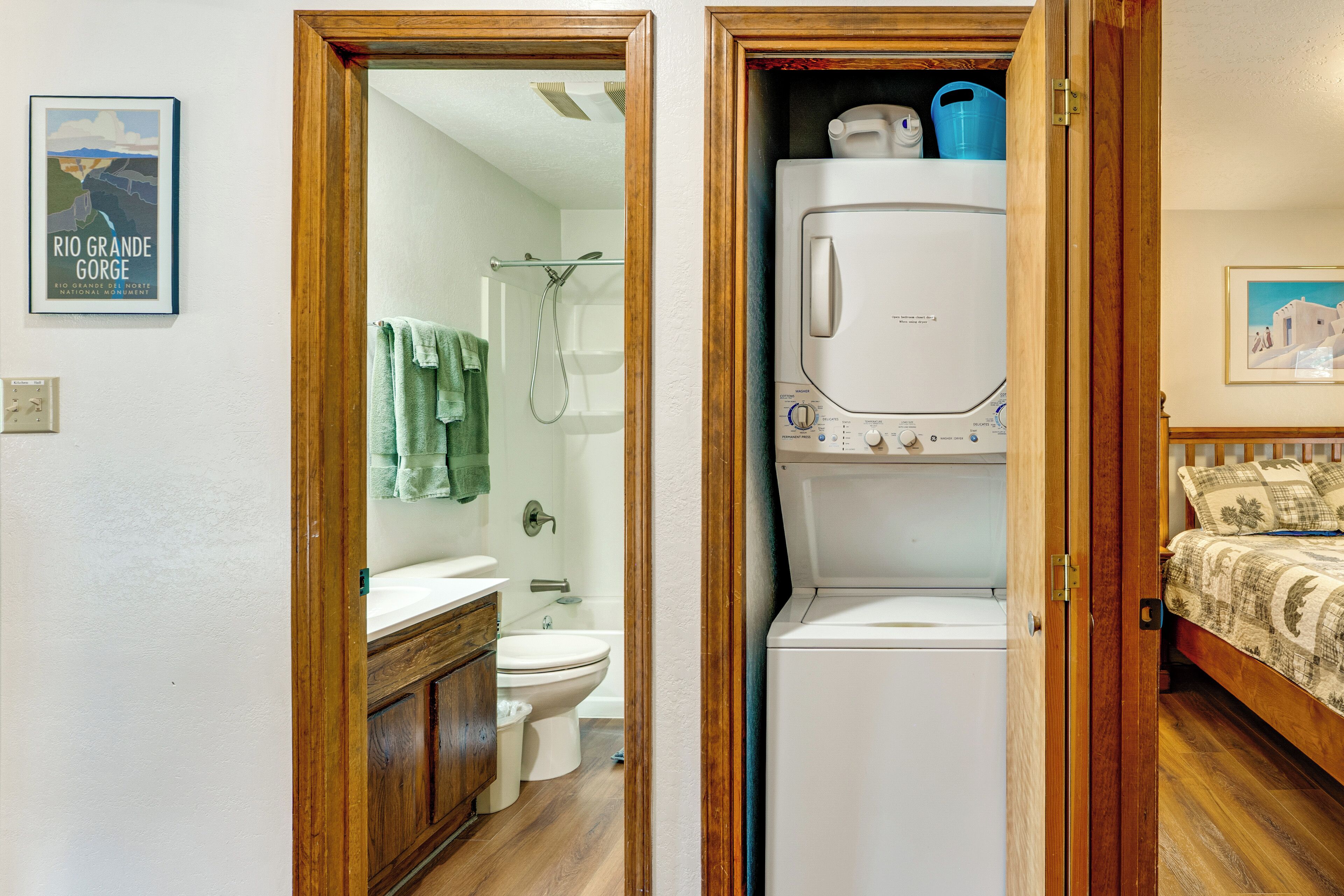 A compact room with a wooden door, a white washing machine, a bed with patterned bedding, a framed picture on the wall, and a bathroom visible through the door.