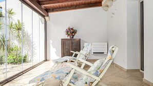 A sunlit room with three sun loungers, a small wooden cabinet with a vase of flowers, and a window overlooking a garden. - Holiday Home 'Villa Fortuna Con Vista Mare' with Sea View, Wi-Fi and Air Conditioning (Baja Sardinia)
