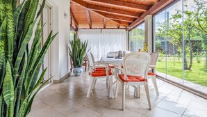 A dining area with a wooden ceiling, white chairs with orange cushions, a white table, and large windows overlooking a garden. - Holiday Home 'Villa Fortuna Con Vista Mare' with Sea View, Wi-Fi and Air Conditioning (Baja Sardinia)