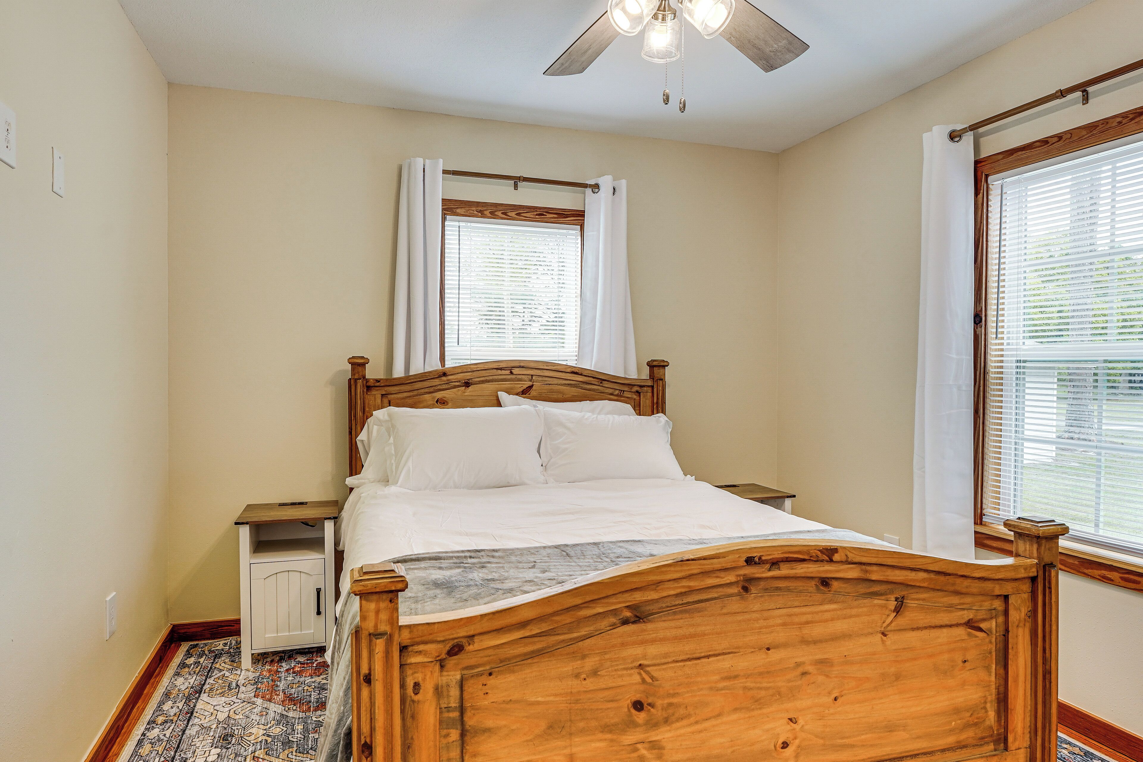 A bedroom with a wooden bed, a nightstand, a small cabinet, a ceiling fan, and a window with blinds.