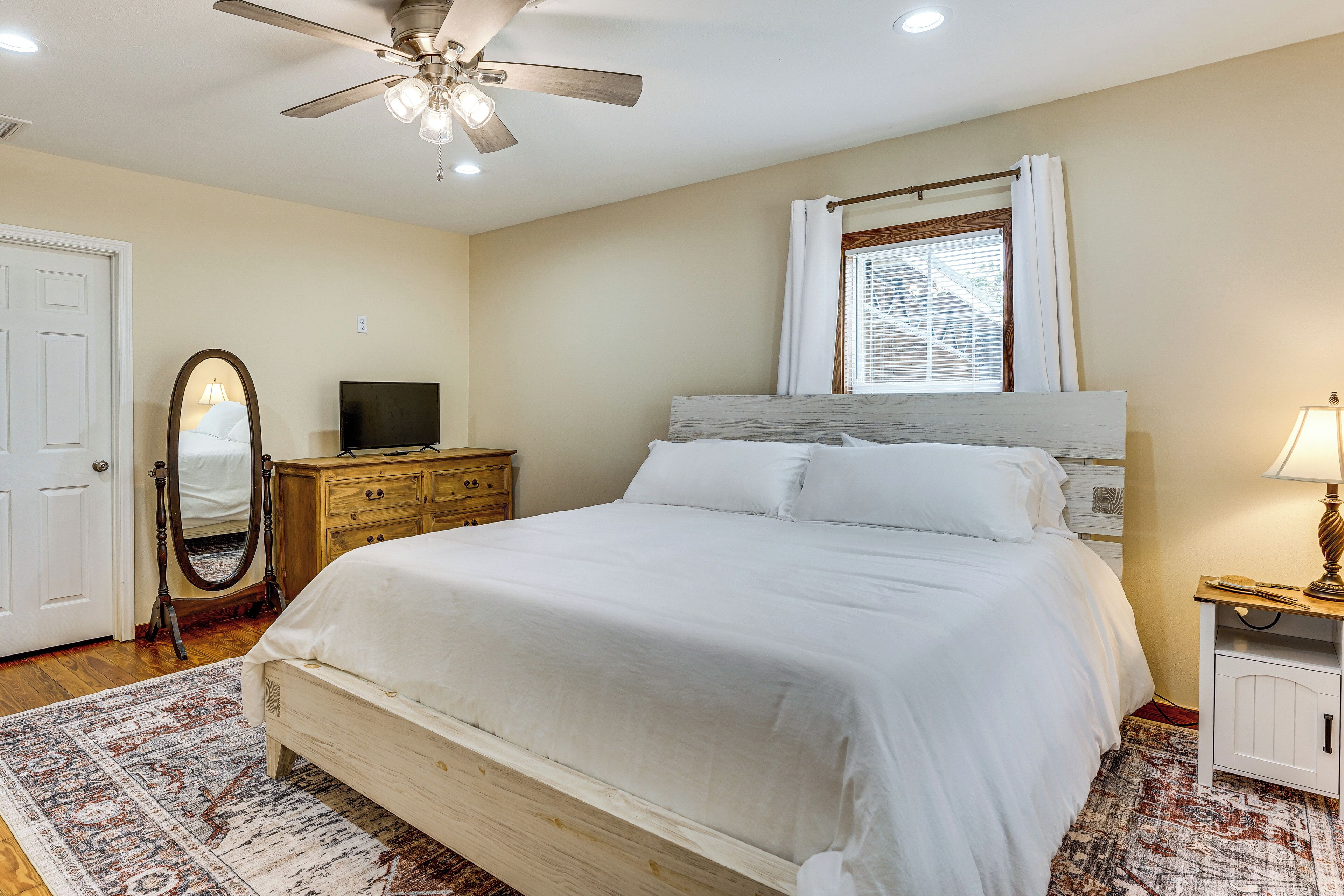 A bedroom with a bed, a ceiling fan, a TV, a dresser, a mirror, and a window with curtains.