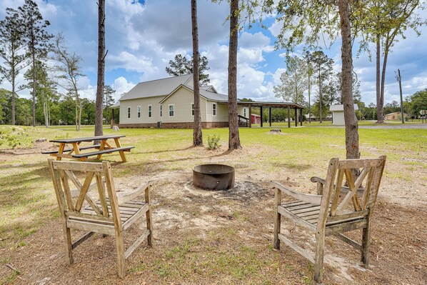 A rustic outdoor seating area with a picnic table and two wooden chairs, a fire pit, and a house in the background.