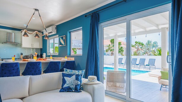 Ein modernes Wohnzimmer mit einer Couch, einem Essbereich mit Stühlen, einer Küche mit Schränken und Blick auf einen Pool sowie Gartenmöbel.