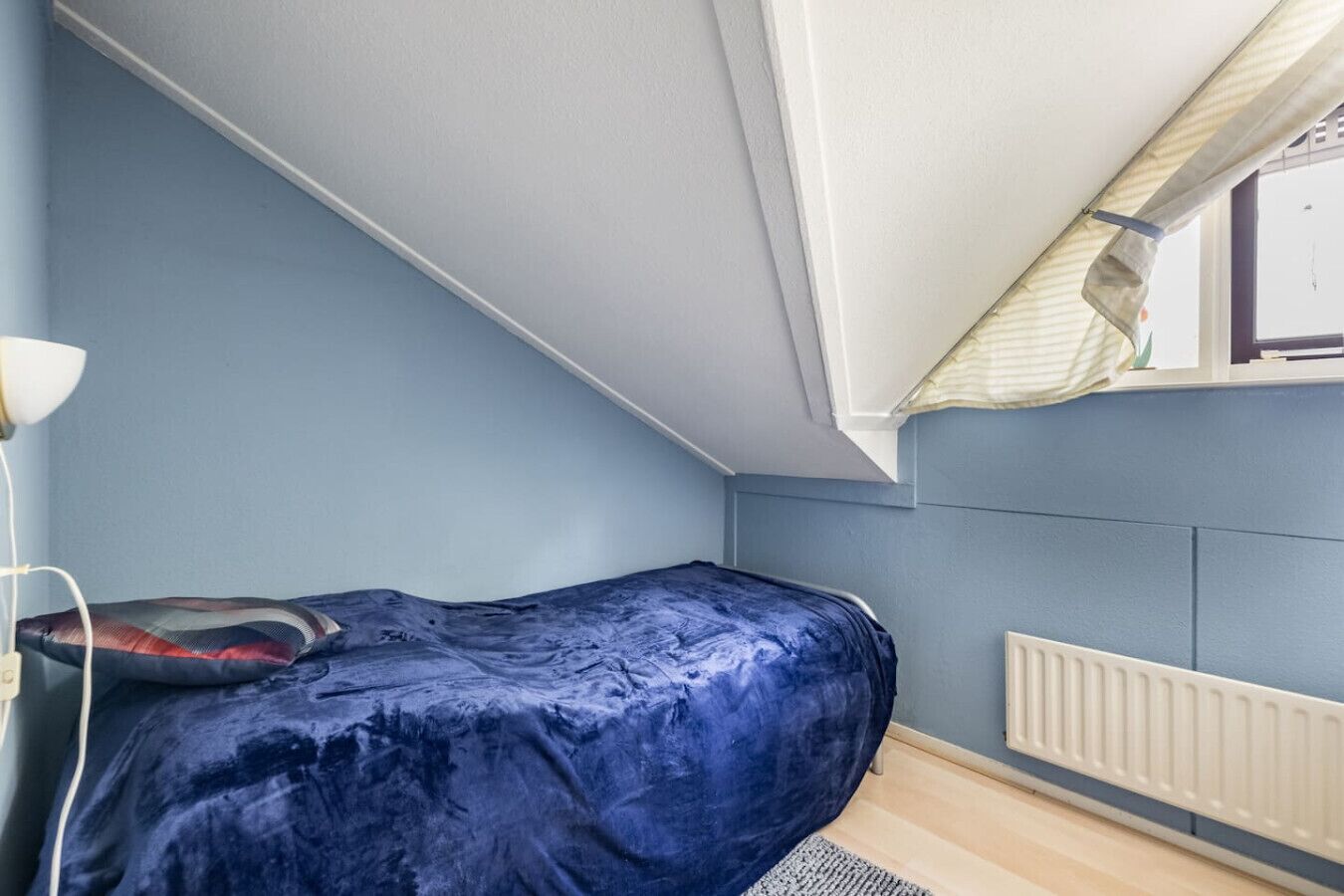A small, single-bed room with a sloped ceiling, a radiator, and a window with blinds.
