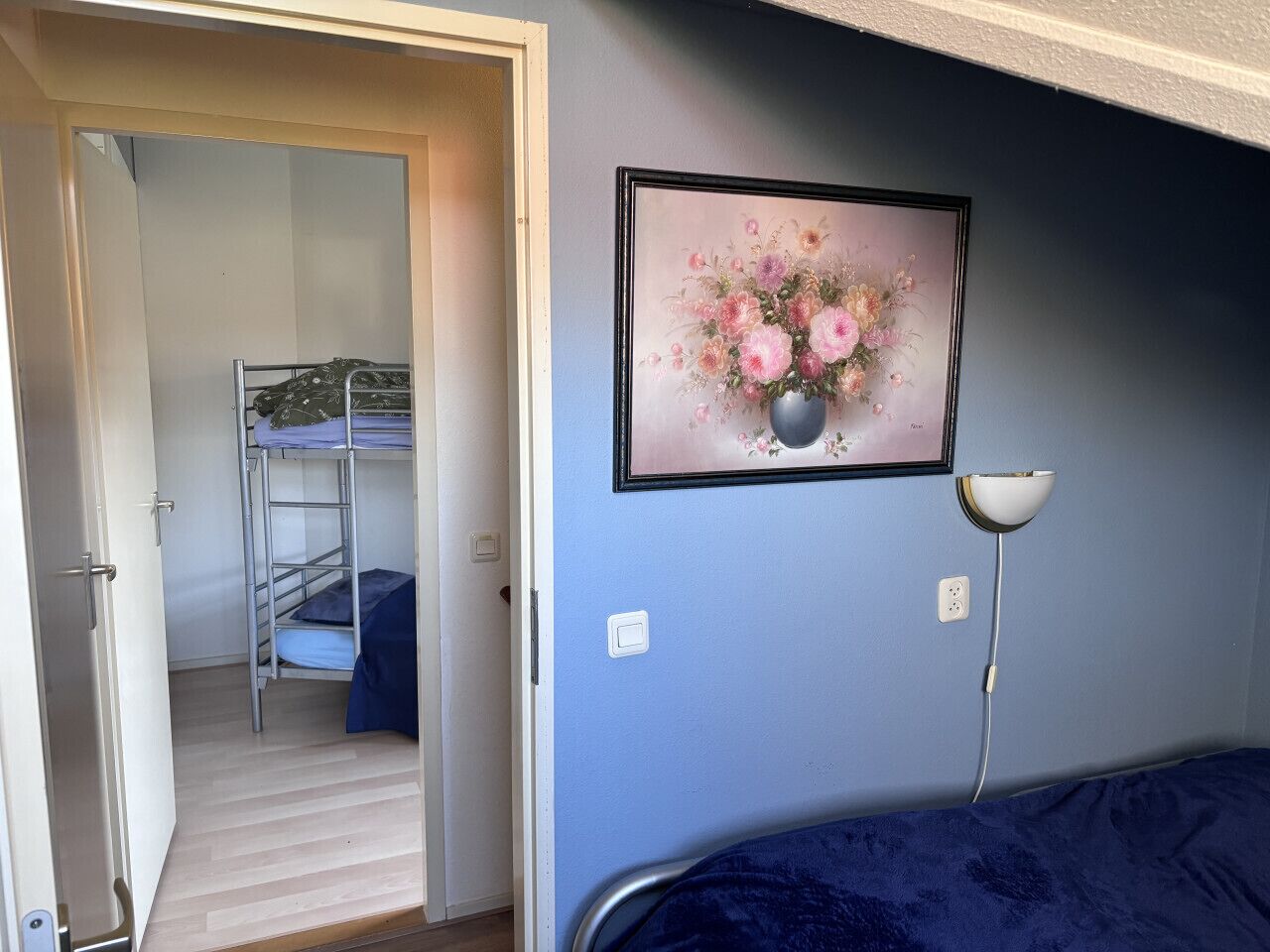 A room with a bunk bed, a framed floral artwork on the wall, a bedside lamp, and a door leading to another room.
