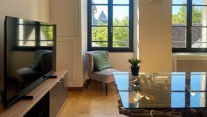 A modern living room with a flat-screen TV, a glass coffee table, a patterned armchair, and a view of a building through a large window. - Appartement du Châtel (Provins)
