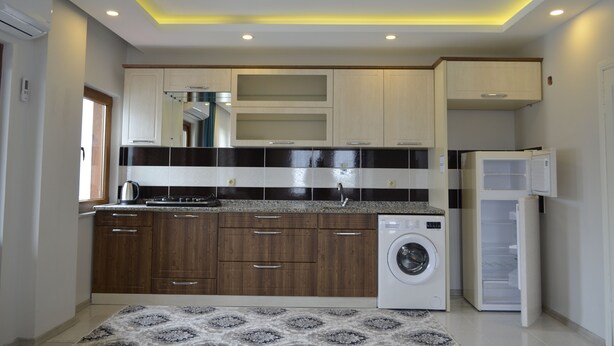 A compact kitchen with wooden cabinets, a built-in washing machine, and a refrigerator. - Özkan Pansiyon (Caykara)