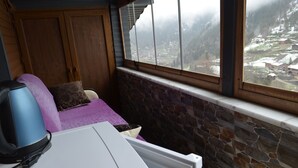 A cozy room with a stone wall, a window with a view of a snowy village, a purple sofa, and a white table with a kettle. - Özkan Pansiyon (Caykara)