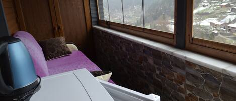 A cozy room with a stone wall, a window with a view of a snowy village, a purple sofa, and a white table with a kettle.