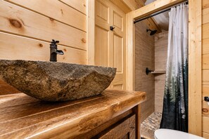 Et interiør af en træhytte med en vask i stentøjslook, træskabe og et bruseområde med et gardin.