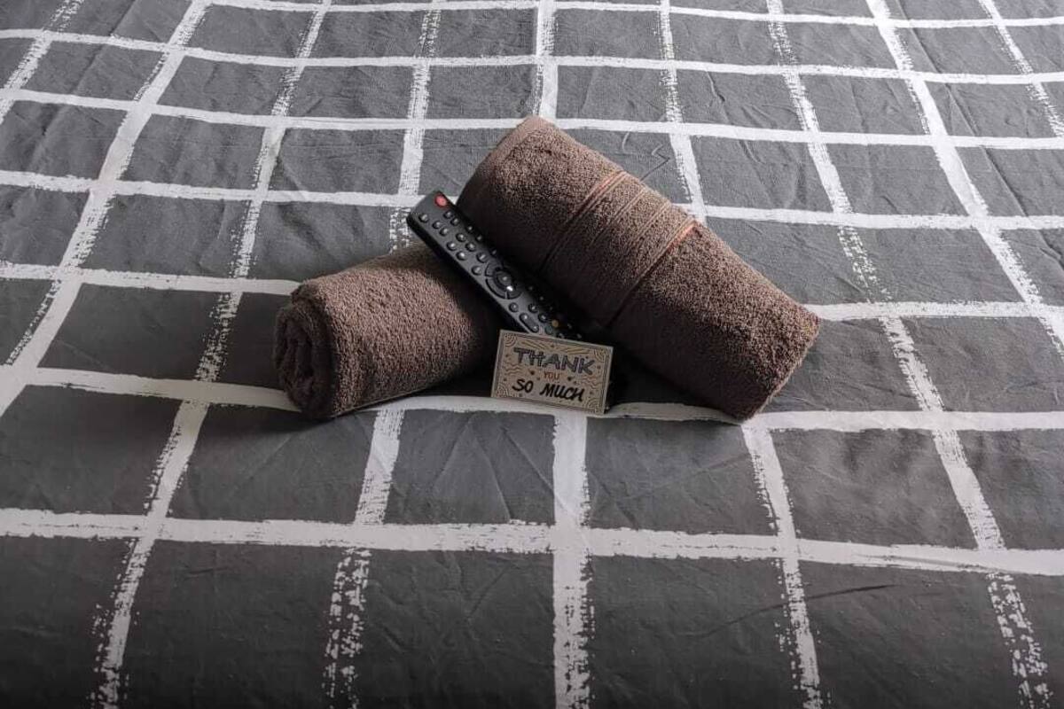 A bed with a checkered bedspread, two pillows, a rolled towel, and a thank you card.