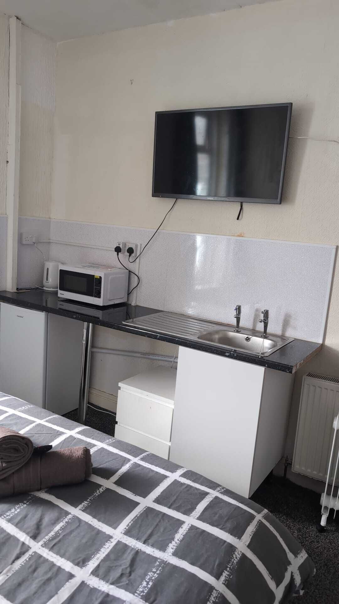 A compact hotel room with a kitchenette, a bed with a checkered blanket, a flat-screen TV mounted on the wall, and a radiator.