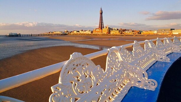 A beach with a pier extending into the water, a lighthouse in the background, and ornate white railings along the shore. - The Galloway Hotel  (Blackpool)