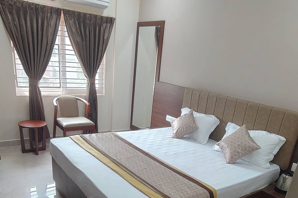A hotel room with a large bed, a chair, a small table, an air conditioning unit, and a mirror.